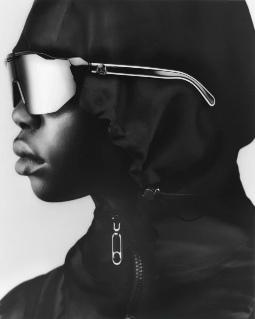Black and white close-up of a person with dark skin, wearing sunglasses and a black hooded jacket, creating a fashion editorial shot.