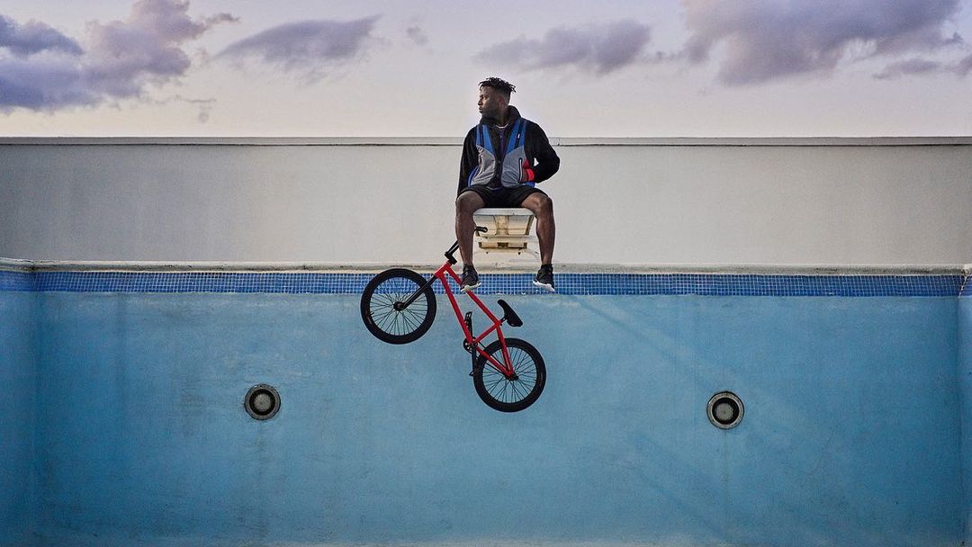Adult man sitting on a white chair with a red BMX bike in front of an empty blue tiled swimming pool with white walls.