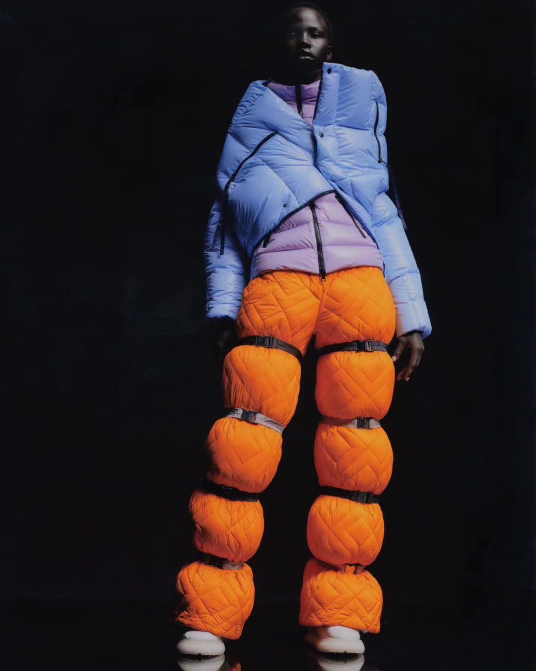 Fashion shot of an adult Black model wearing an avant-garde puffer jacket and orange puffer pants against a dark studio backdrop.