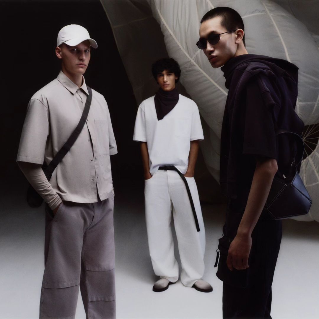 Three fashion models pose in a photo studio. One wears a light beige outfit with a hat and bag, another is in white, and one is in black.