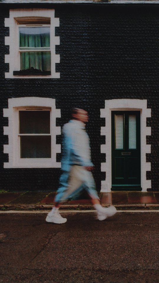A blurry image of an adult walking along a sidewalk in front of a dark building with white trim around windows and doors.