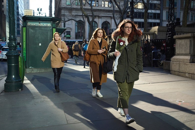 Three women walk on a sunlit city sidewalk near Bryant Park, wearing coats and carrying items on a clear day, in an urban setting.