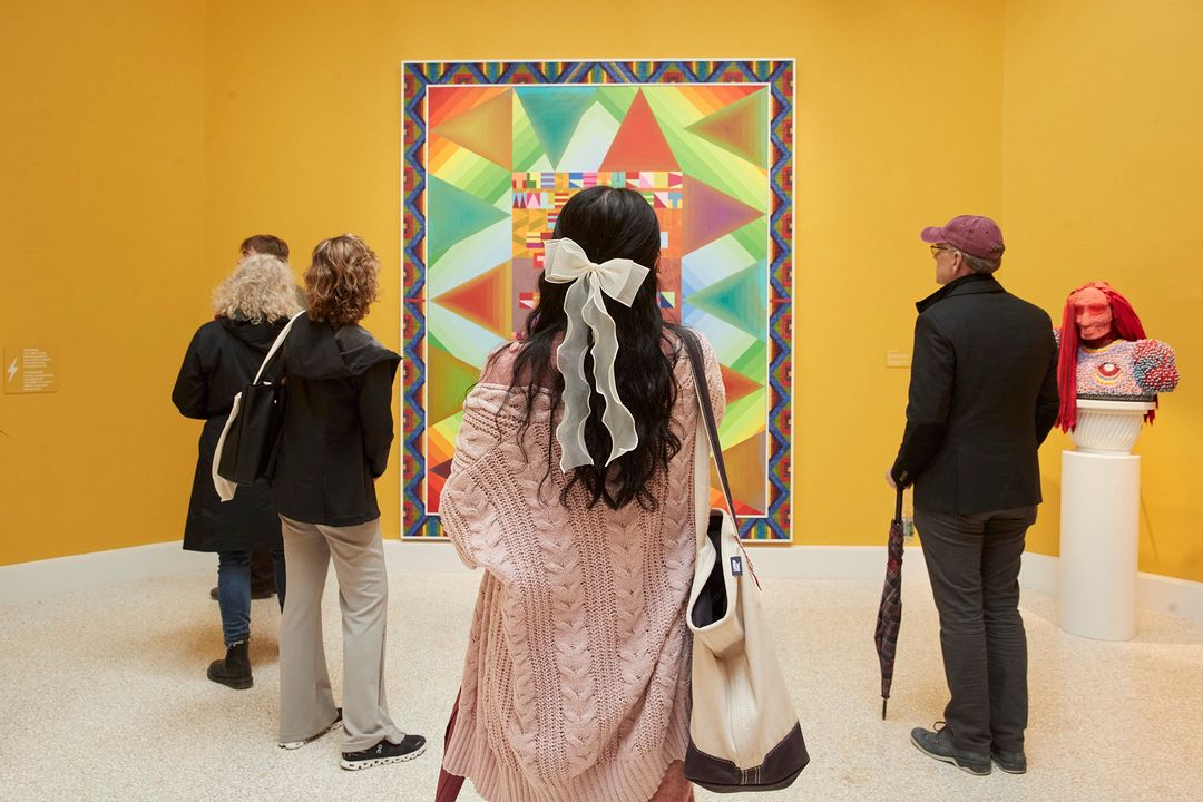 People view a colourful geometric art exhibit inside a museum. The abstract artwork is framed in a gallery against a yellow wall.