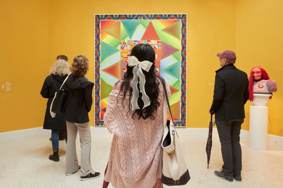 People view a colourful geometric art exhibit inside a museum. The abstract artwork is framed in a gallery against a yellow wall.