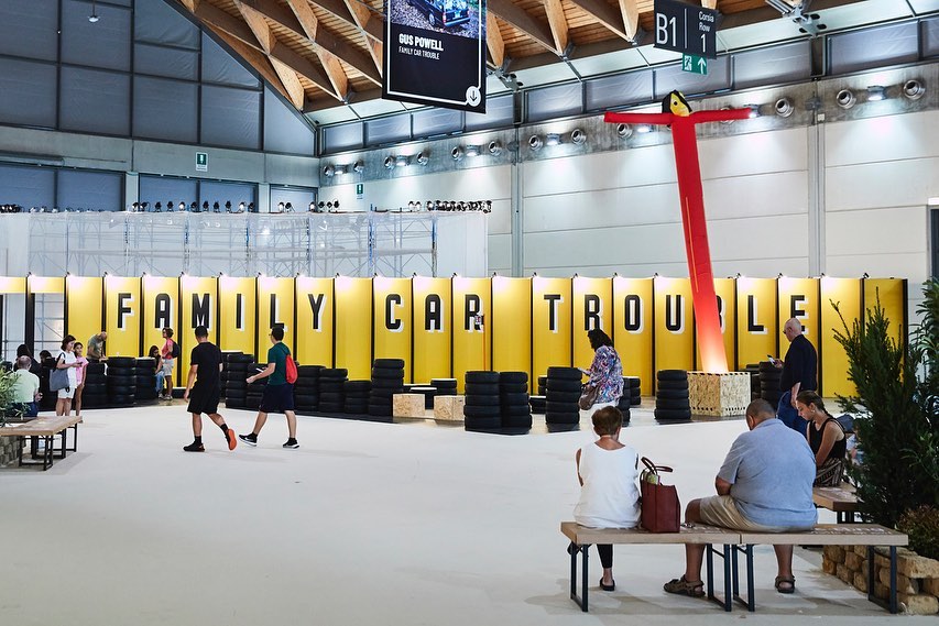Large indoor exhibition hall featuring yellow backdrops displaying the text 'FAMILY CAR TROUBLE,' groups of people, and stacked tires.