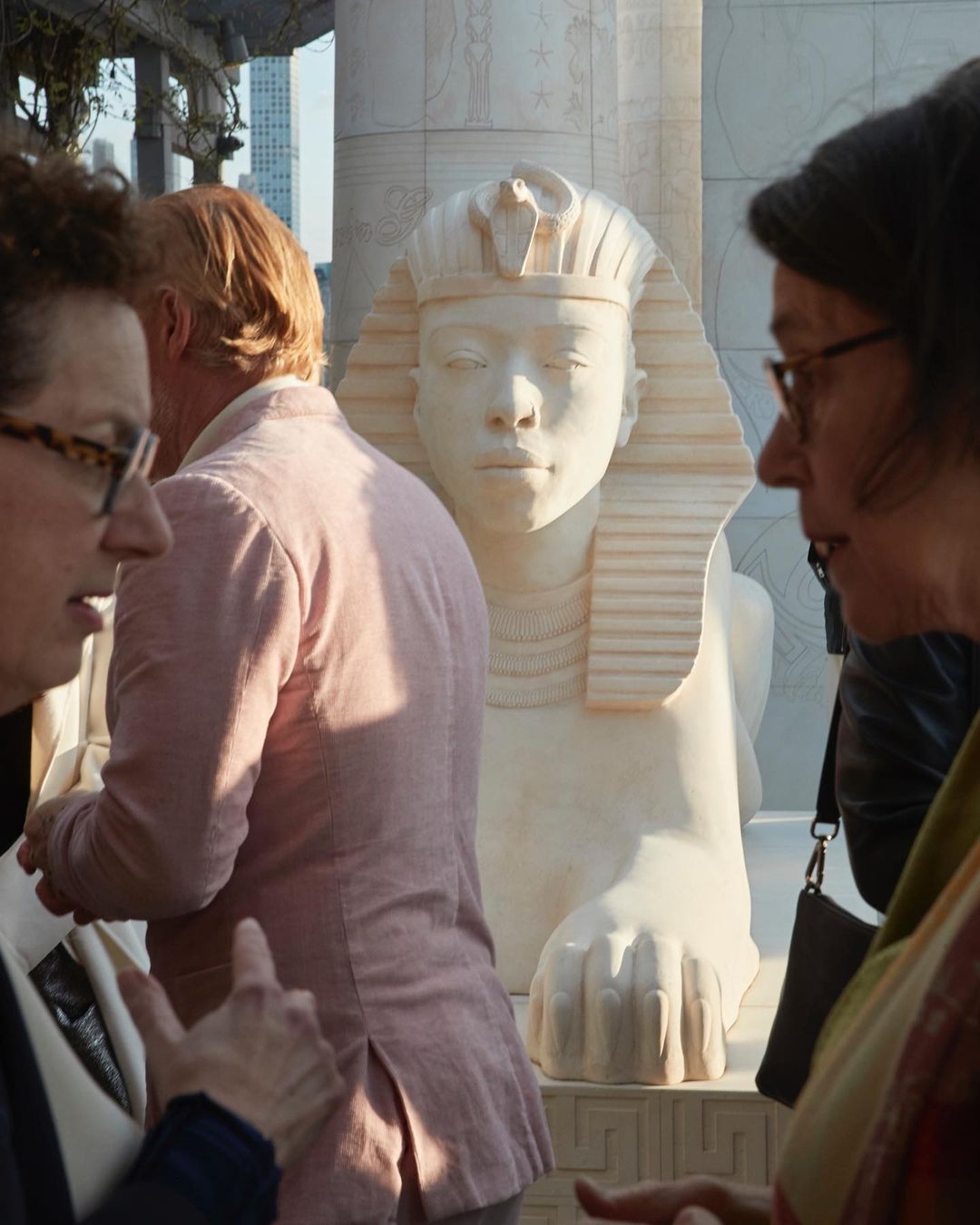 Outdoor scene with two people in the foreground discussing an Egyptian sphinx sculpture standing on a stone pedestal in the background.