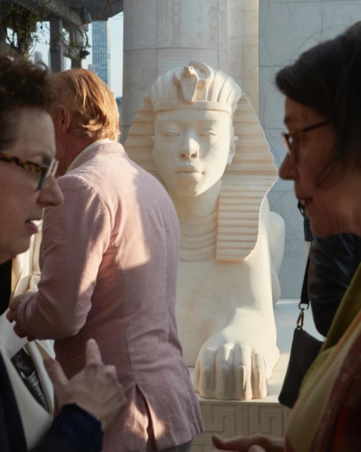 Outdoor scene with two people in the foreground discussing an Egyptian sphinx sculpture standing on a stone pedestal in the background.
