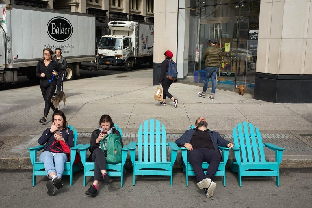 People relax in turquoise chairs on a city street with pedestrians and delivery trucks in the background on a bright day.