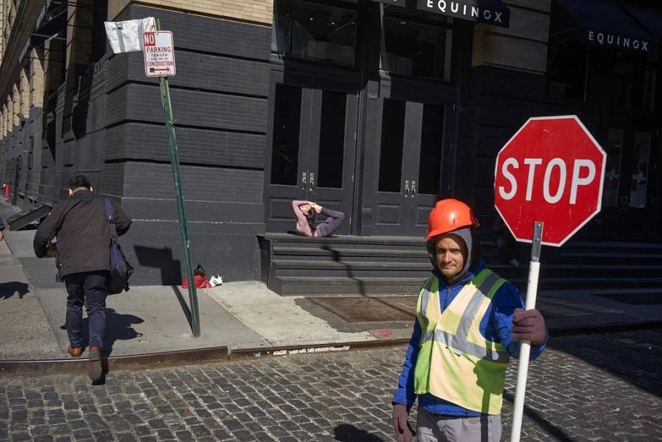 Street scene: Construction worker holds a stop sign; a woman in yoga pose sits on building steps; a man walks past on cobblestone street.