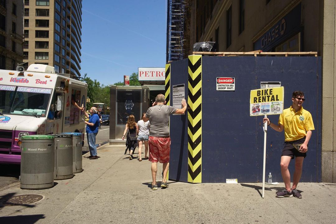 Outdoor scene in New York City shows a bike rental promoter, people walking, an ice cream truck, construction barriers, and city infrastructure.