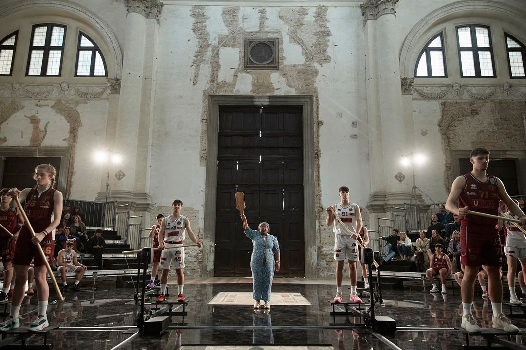 Basketball players stand on machines as a woman in a blue dress holds up a wooden object in an old building with spectators.