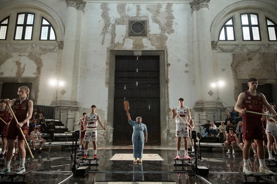 Basketball players stand on machines as a woman in a blue dress holds up a wooden object in an old building with spectators.