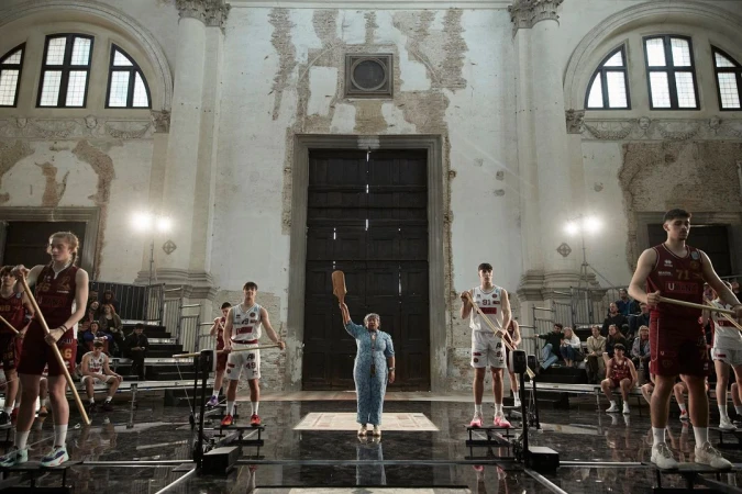 Basketball players stand on machines as a woman in a blue dress holds up a wooden object in an old building with spectators.