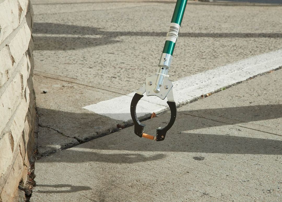 Close-up of a litter picker tool holding a cigarette butt on a concrete surface with a brick wall in the background on a sunny day.