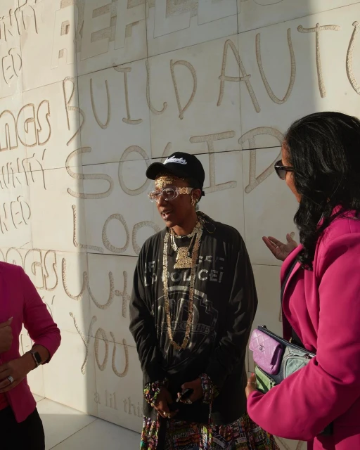 Two women outdoors in front of wall with carved text; woman in streetwear style and gold jewelry is primary subject.