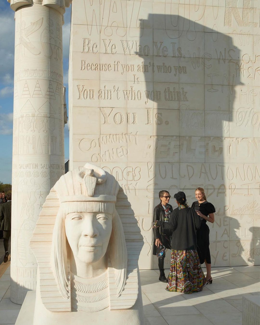 Outdoor shot captures a stone sphinx sculpture with carved text behind, and three women in the background on a bright day.