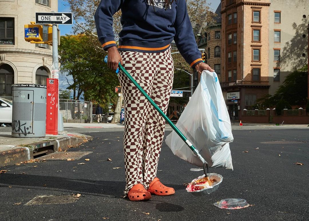 Adult picks up trash with a trash grabber on a street corner near buildings and a traffic light on a sunny day.