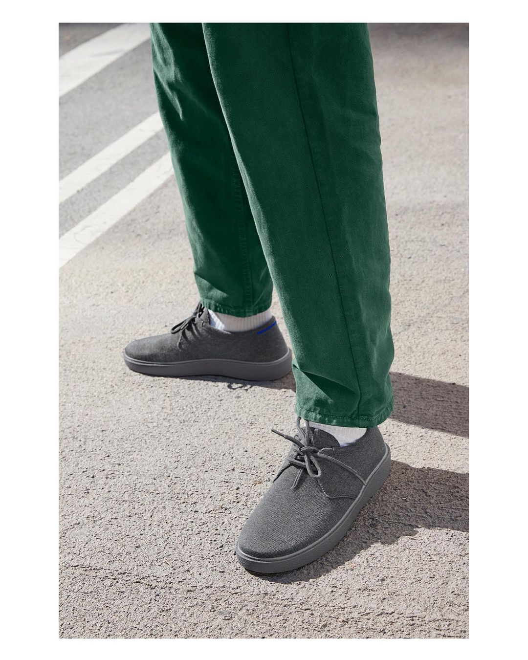 Person wearing gray shoes and green pants stands on gray pavement, footwear and fashion concept, daytime outdoor setting.