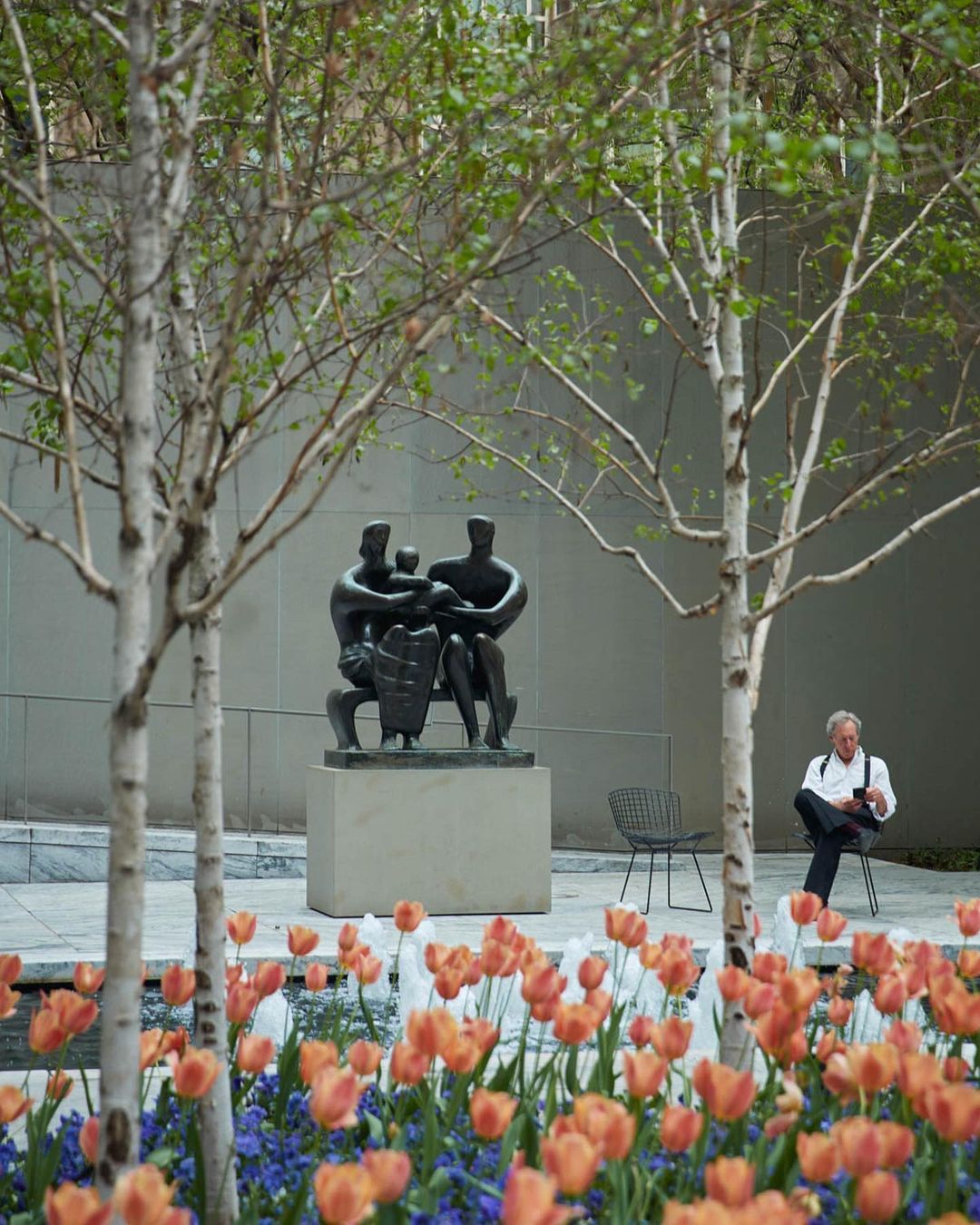 Bronze sculpture of a family group in a city garden, with a seated man in the background and vibrant tulips and trees in the foreground.