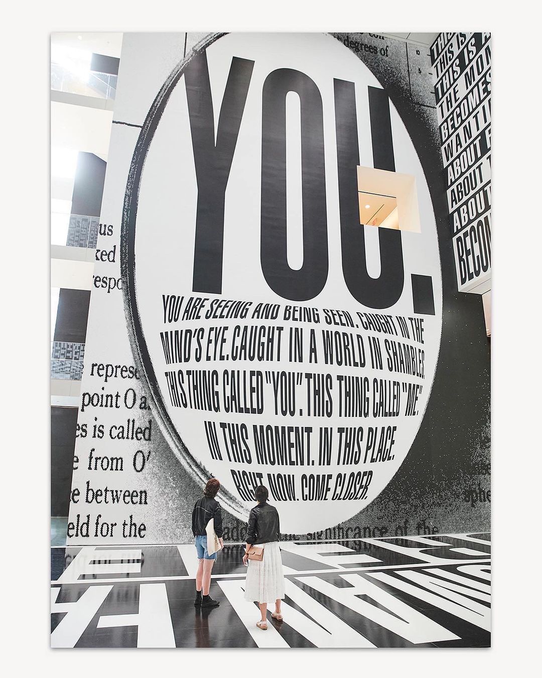 Two people view a large-scale typographic art installation indoors, featuring overlaid black and white text, and black and white flooring.