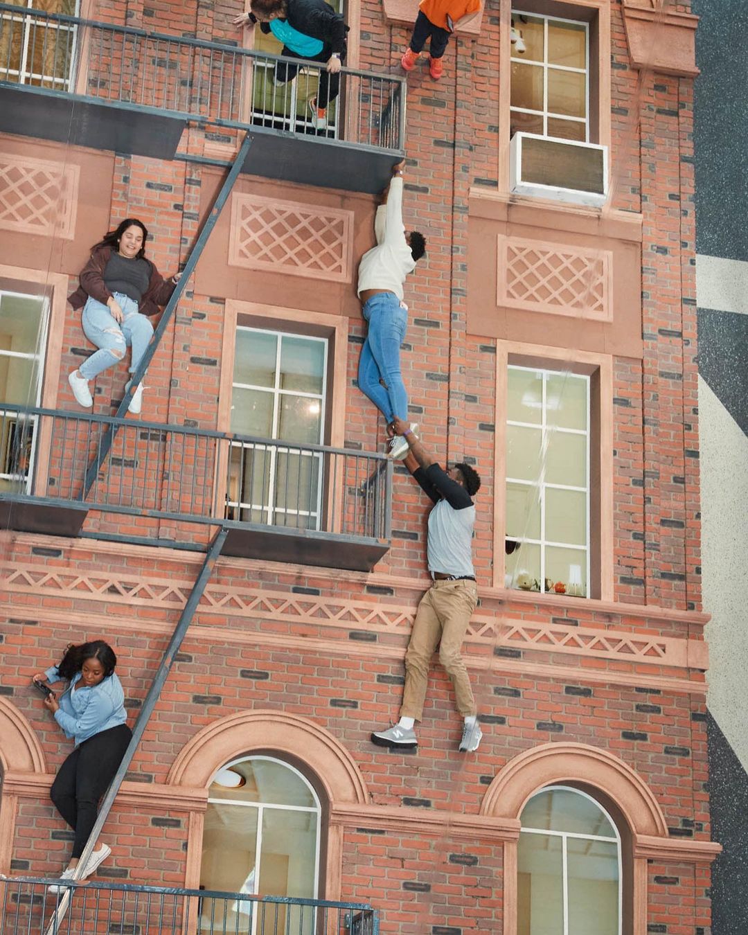 Brick building with fire escape featuring six people in various poses of climbing or descending, shot from a low angle in daylight.