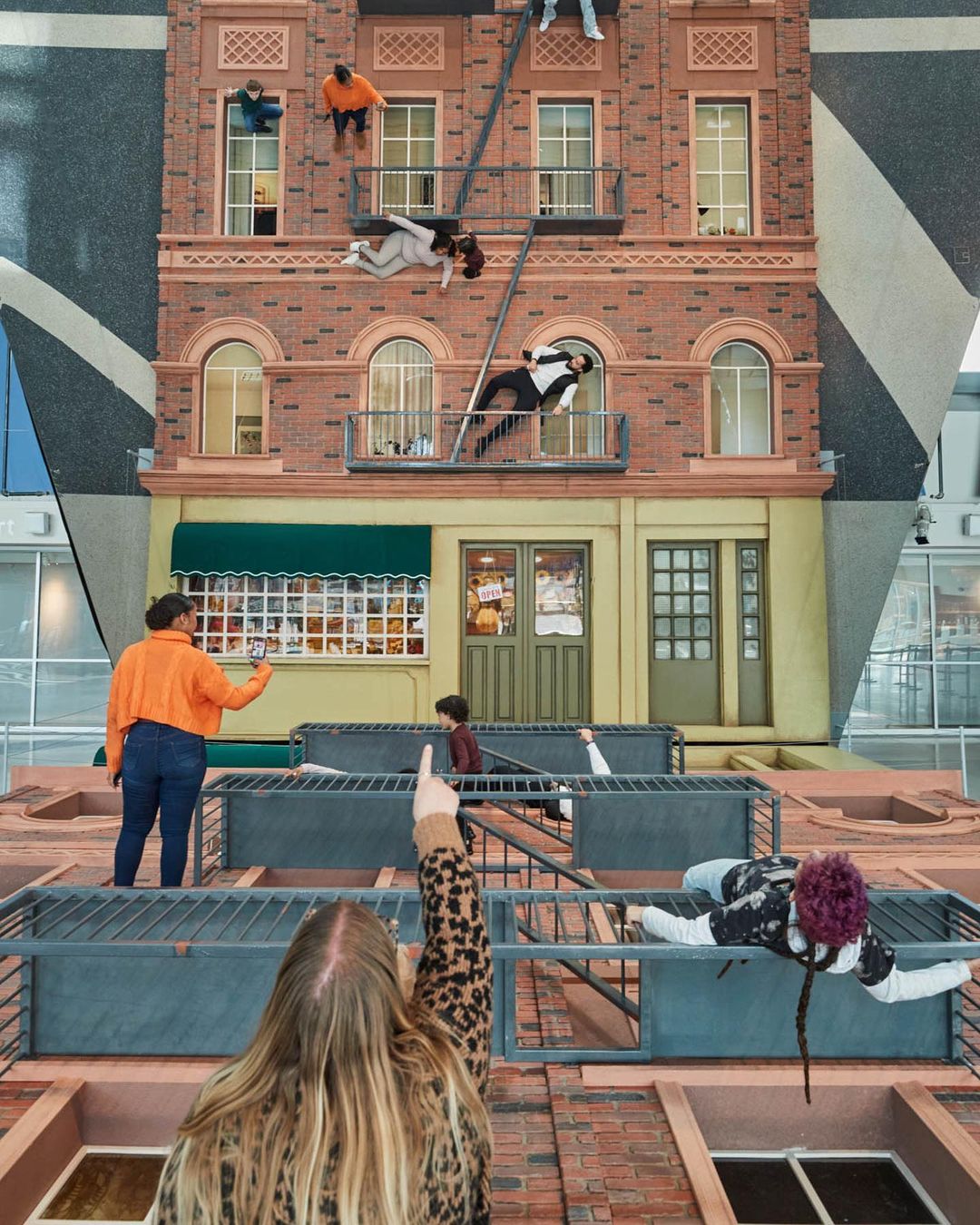 Art installation featuring figures posing on a realistic-looking building facade inside a modern space, observed by people from below.