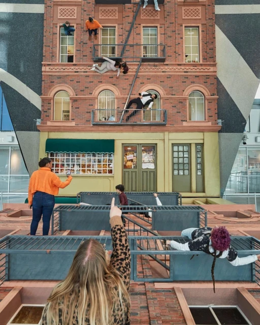 Art installation featuring figures posing on a realistic-looking building facade inside a modern space, observed by people from below.