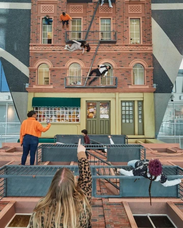 Art installation featuring figures posing on a realistic-looking building facade inside a modern space, observed by people from below.