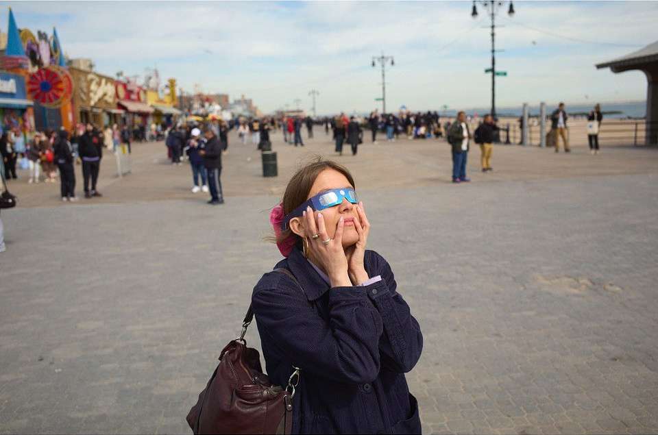 Woman wearing solar eclipse glasses looks up during the daytime at a boardwalk with many people in the background.
