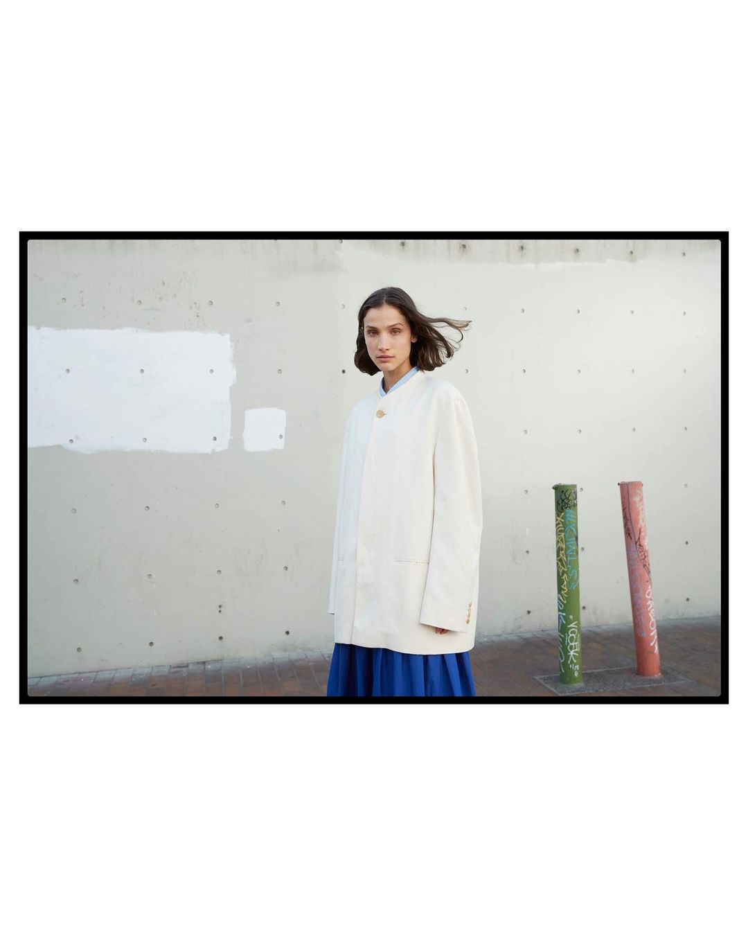 Woman in a white jacket and blue skirt stands in an outdoor setting with a concrete wall and painted pipes in the background.