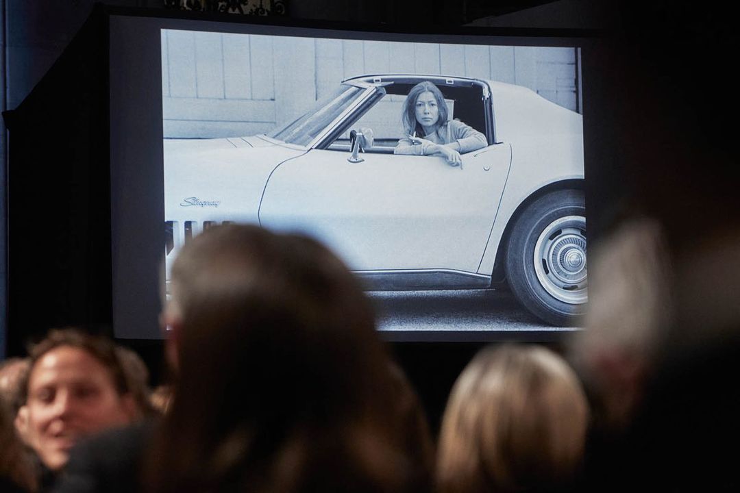 Woman sits inside a white classic Stingray car, visible on a large projector screen for an audience in a darkened indoor venue.