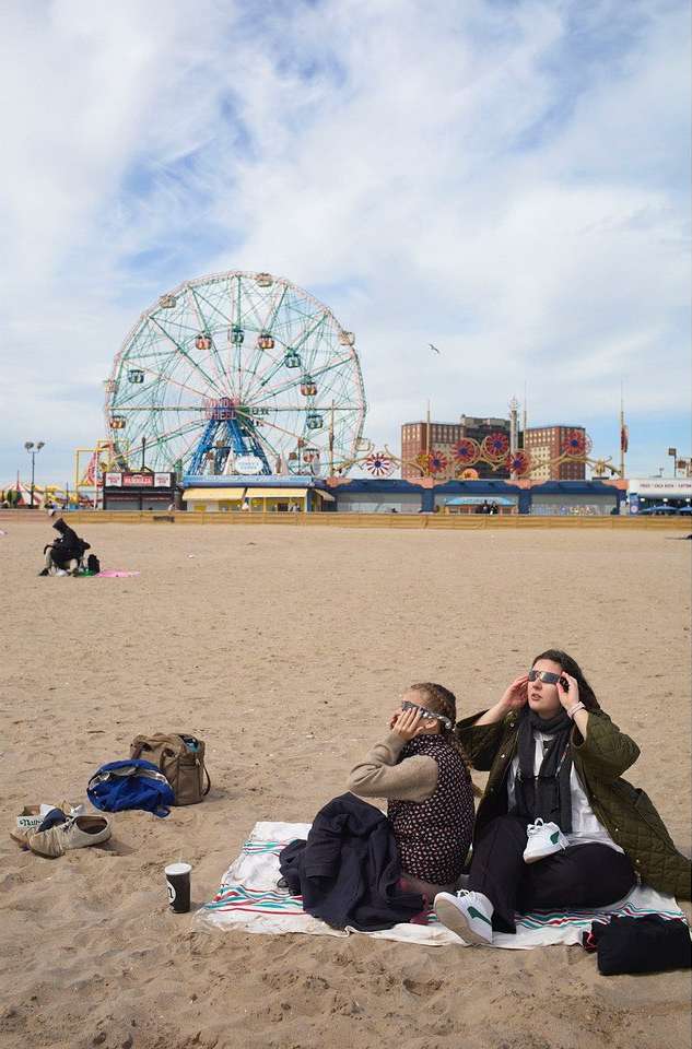 Two women sit on a beach with a Ferris wheel in the background, shielding their eyes from the sun, with bags and personal items.
