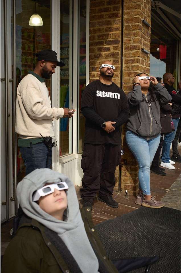 Several people wearing protective glasses look upwards, standing on a sidewalk next to a brick building on a sunny day.