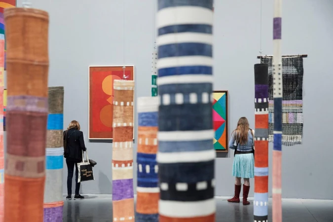 Two women at art gallery viewing colorful hanging textile art and framed geometric paintings in a brightly lit indoor exhibit space.