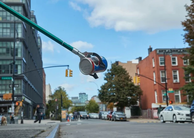 Close-up of a trash grabber tool holding an empty soda can with city street, buildings, and blue sky with clouds in the background.