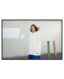 Woman wearing a cream jacket and blue skirt standing outdoors in front of a concrete wall with two painted bollards.