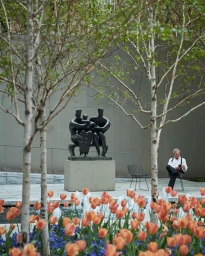 Man seated on chair in a sculpture garden behind orange tulips and birch trees by Gus Powell