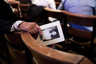 Close-up of a person holding a memorial service program featuring a black-and-white portrait, seated in a wooden chair.