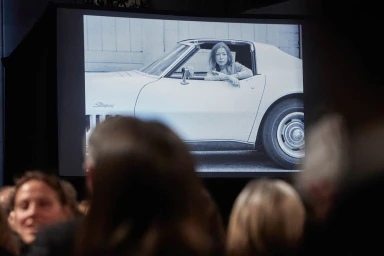 Monochrome projection of a woman sitting in a sports car viewed above an audience in a darkened room