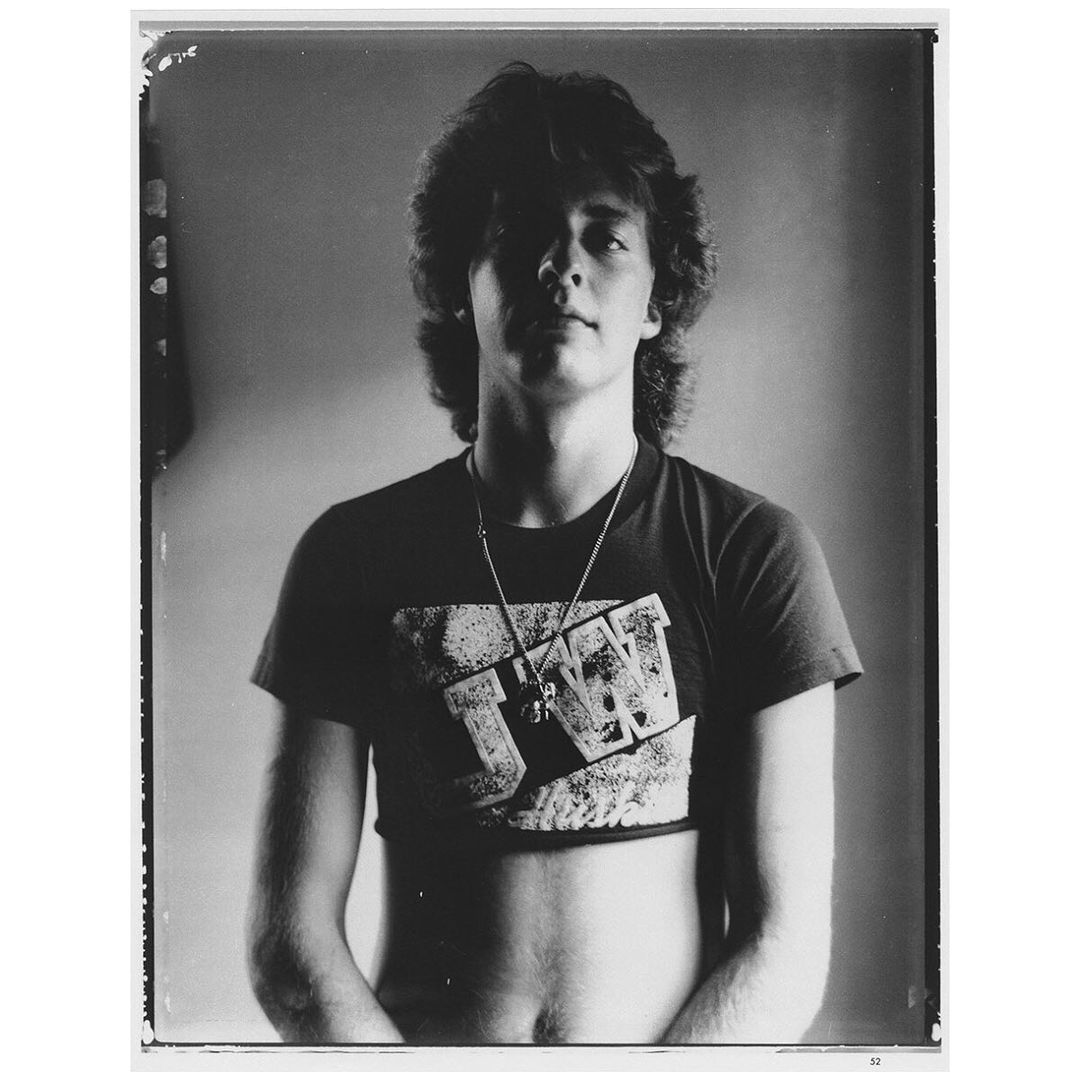 Monochrome portrait of a shirtless adult male with a short top, shoulder-length curly hair, and a graphic tee with a pendant necklace.