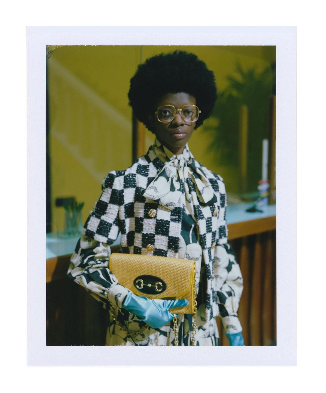 Adult female model in a checkered jacket, gold glasses, and blue gloves holding a gold purse poses in an indoor setting.
