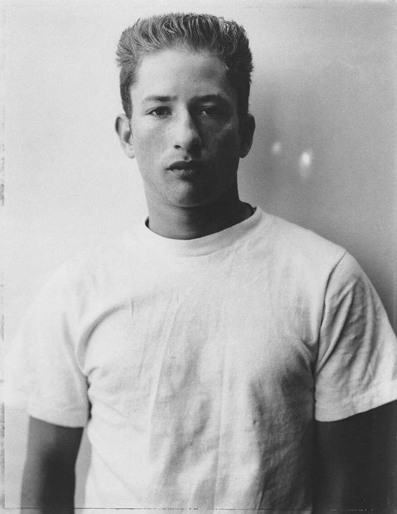 Monochrome portrait of a young adult male with short, curly hair wearing a plain white t-shirt, standing indoors against a neutral background.