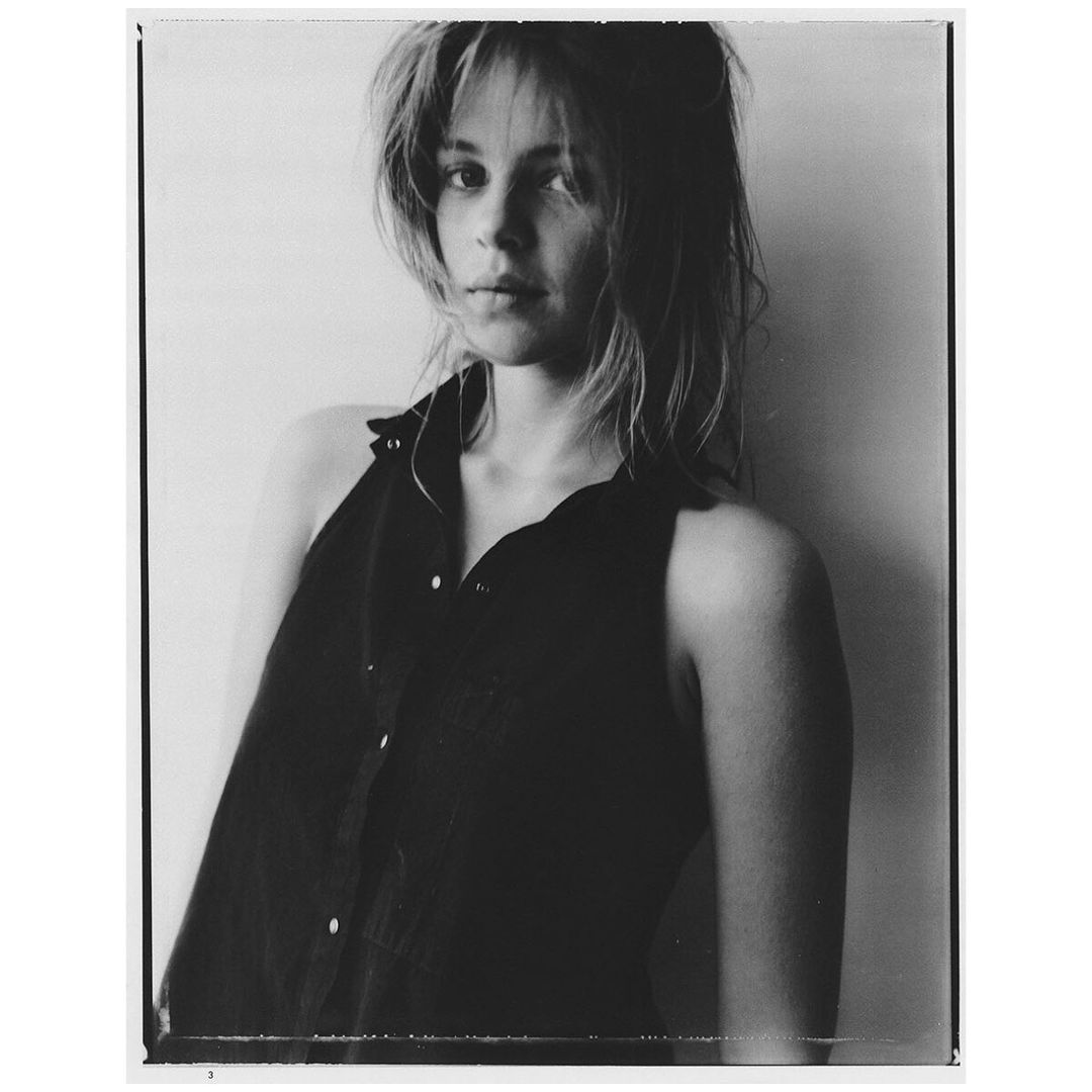 Monochrome portrait of a young woman with shoulder-length hair, wearing a sleeveless button-down shirt, against a plain background.