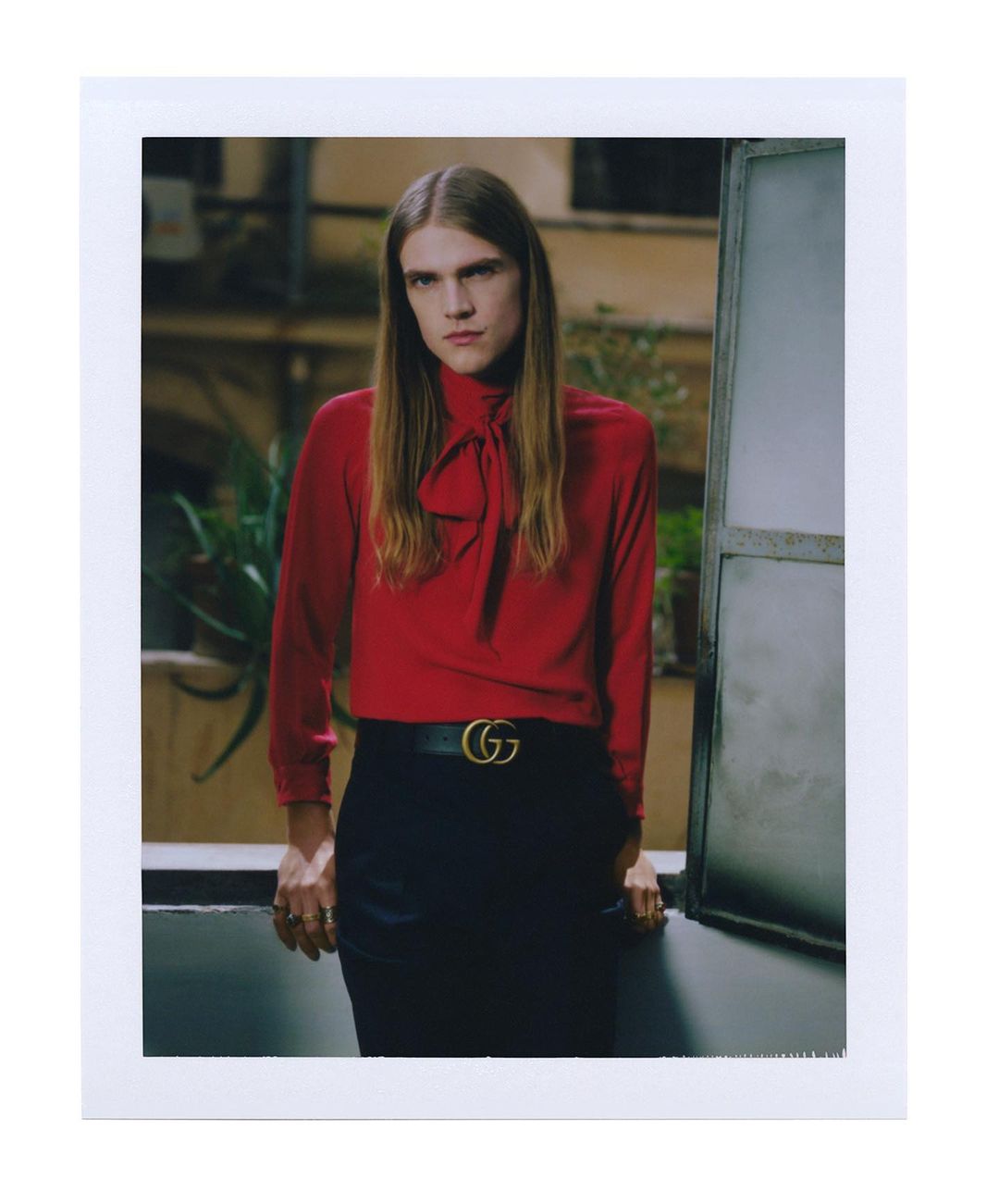 A fair-skinned person with long blonde hair stands in a red blouse and black skirt with a logo belt, posing indoors by a window.