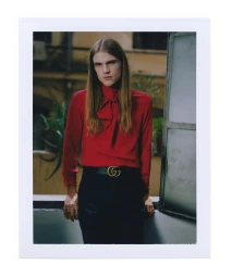 Portrait of a long-haired man in a red pussy-bow blouse and dark trousers with a double G belt, framed in a white polaroid border.