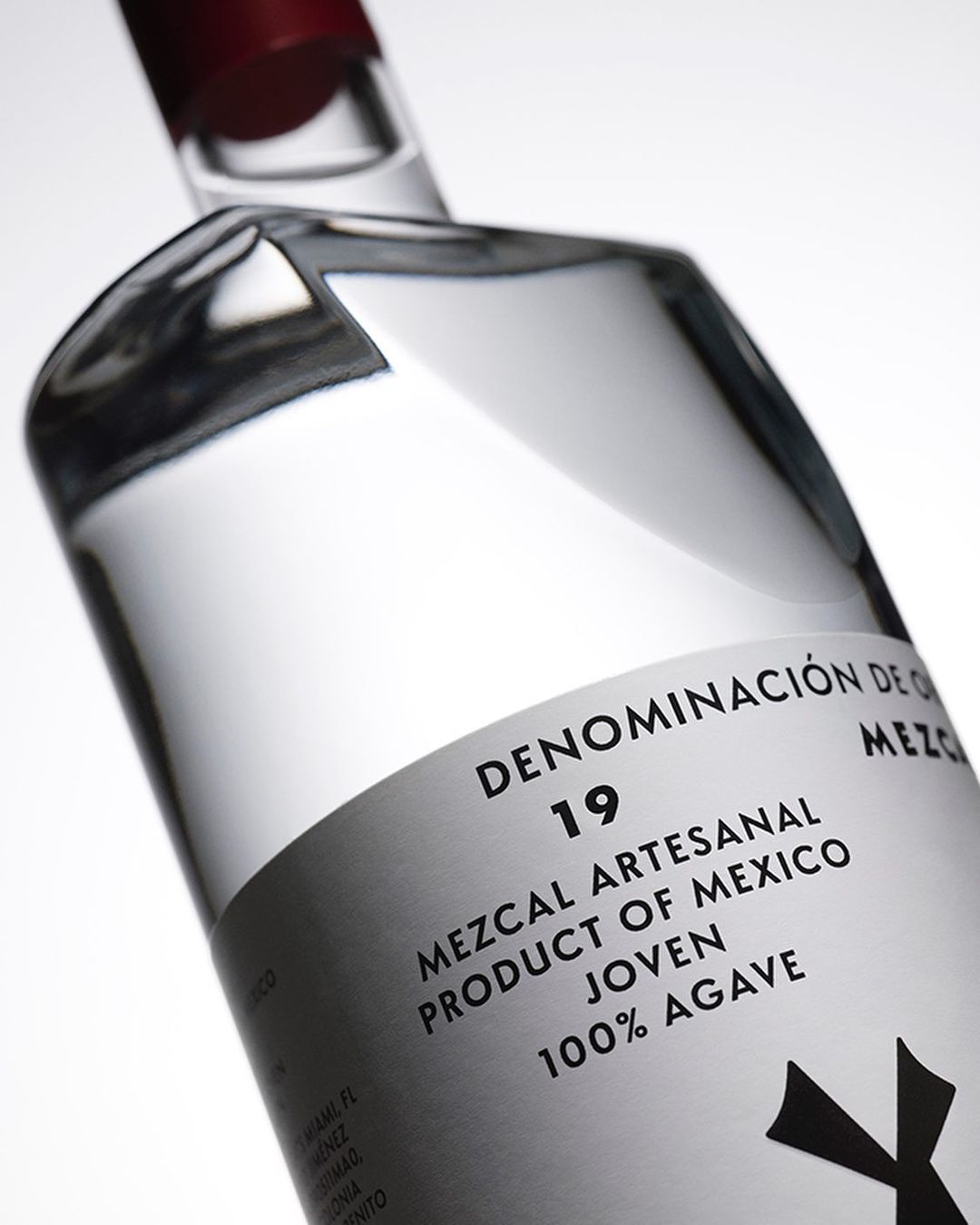 Clear glass mezcal bottle with a red cap, featuring a white label with black text, angled in front of a white backdrop.