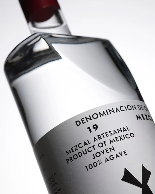 Clear glass mezcal bottle with a red cap, featuring a white label with black text, angled in front of a white backdrop.
