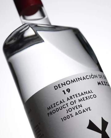 Clear glass mezcal bottle with a red cap, featuring a white label with black text, angled in front of a white backdrop.