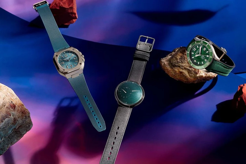 Three luxury wristwatches are displayed against a vibrant backdrop, two resting on rocks, showcasing design and craftsmanship.