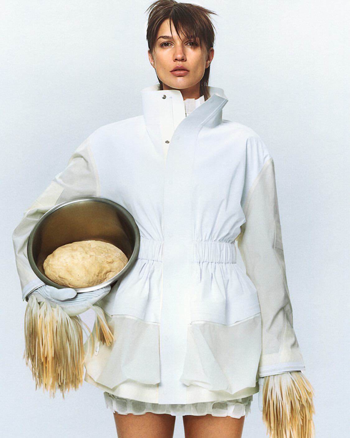 Woman in white jacket holds bowl of dough in studio. Minimalist fashion shot with neutral tones and unusual food styling concept.